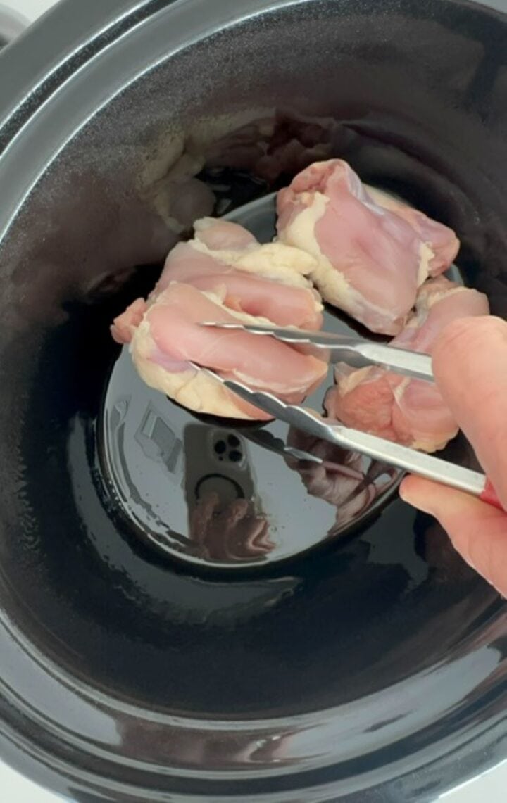 Using aluminum tongs to place pink, raw chicken thighs in a black slow cooker liner.