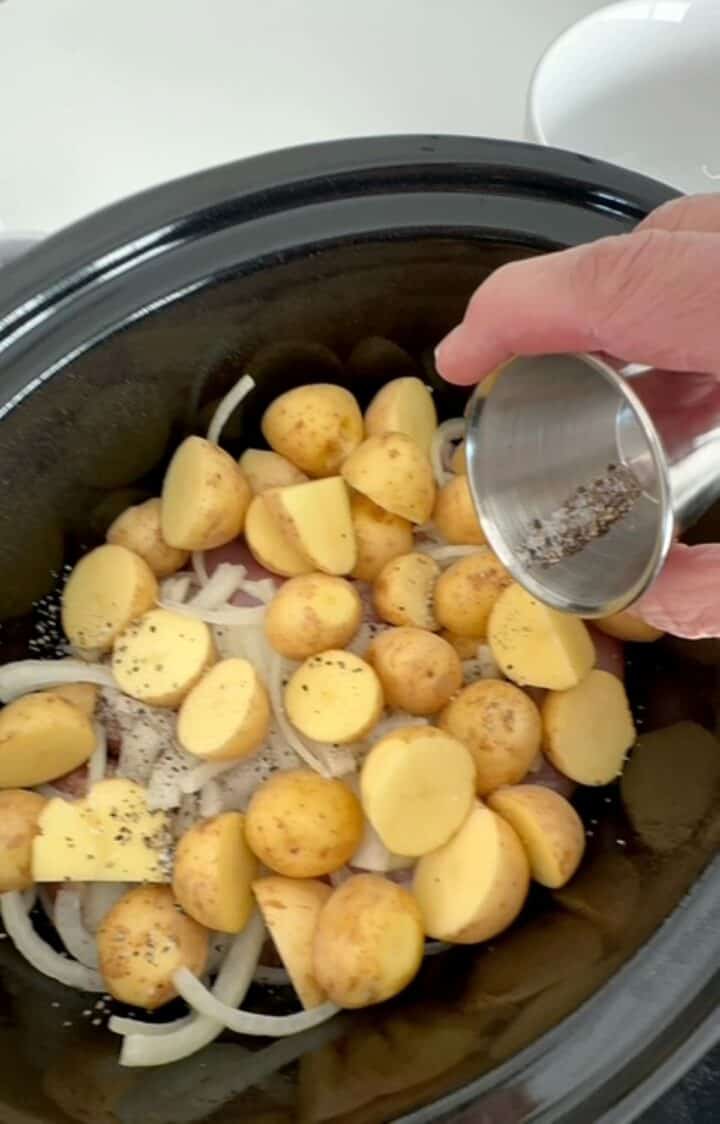 Large crystals of salt and black pepper are sprinkled over yellow baby potatoes and onions in slow cooker.