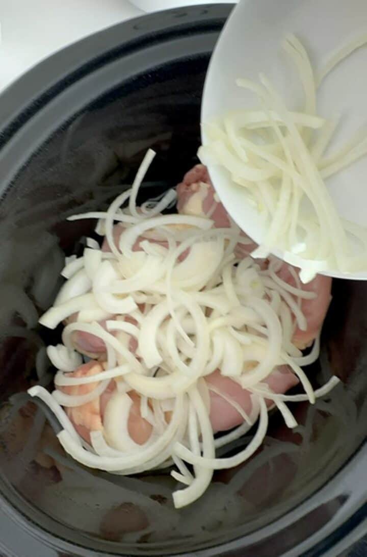 Pouring opaque, white onion slices from a white bowl over pink raw chicken in a black slow cooker.