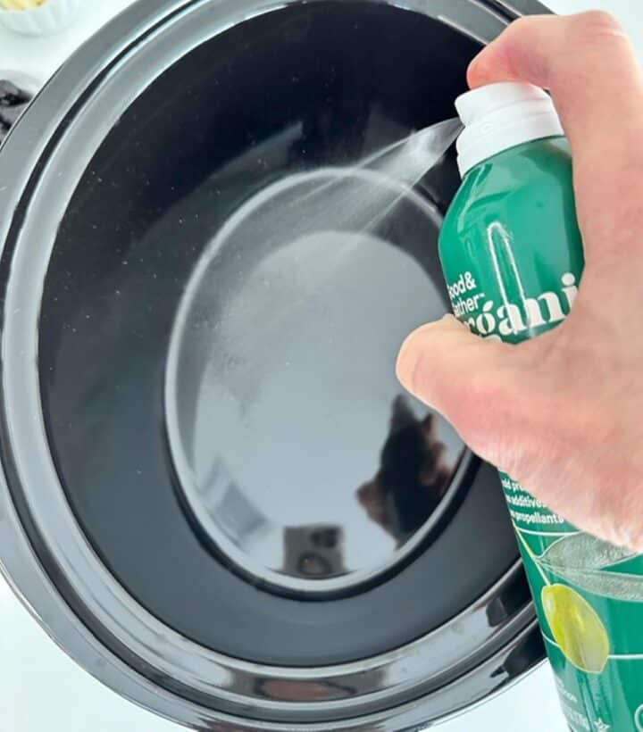 Olive oil spray in a green bottle coats a shiny black slow-cooker liner.