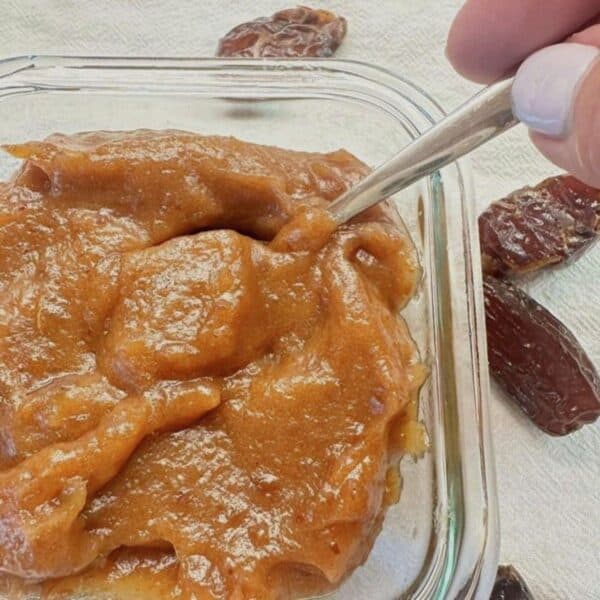 Thick, caramel colored date paste in clear, square dish with a spoon in it.