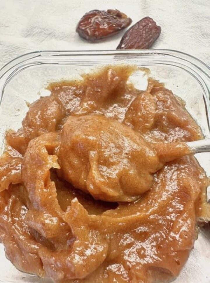 Square glass container of thick caramel colored date paste with a spoon inserted.