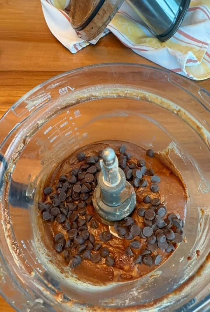 Chocolate chips on top of brown chocolate brownie batter in a food processor bowl.
