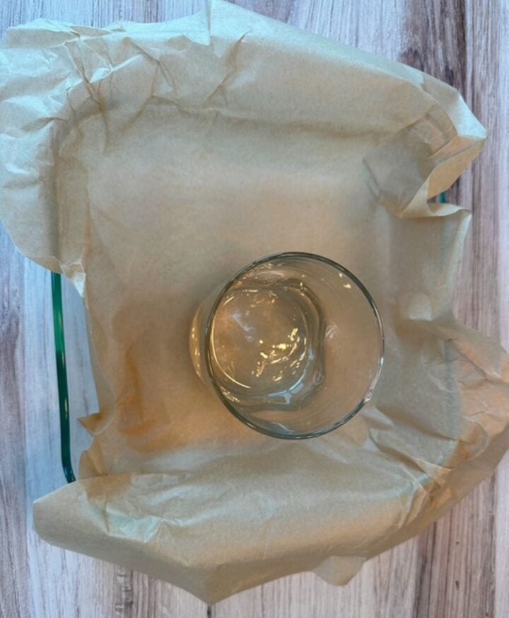 Clear square baking pan contains a brown parchment paper liner with a heavy glass on top of it.