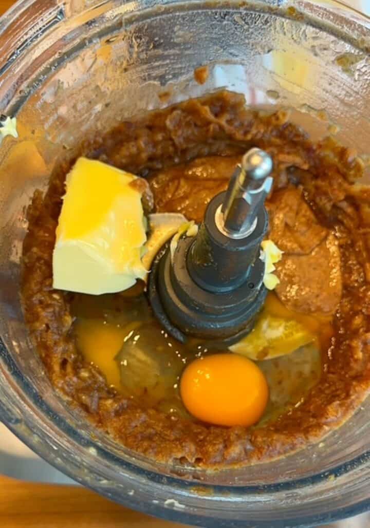 Open food processor holding brown date paste plus a raw egg, butter, and other wet ingredients.