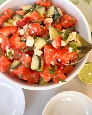Red, juicy watermelon, strawberry bits, and green-white cucumbers as salad in a white bowl with a large spoon.