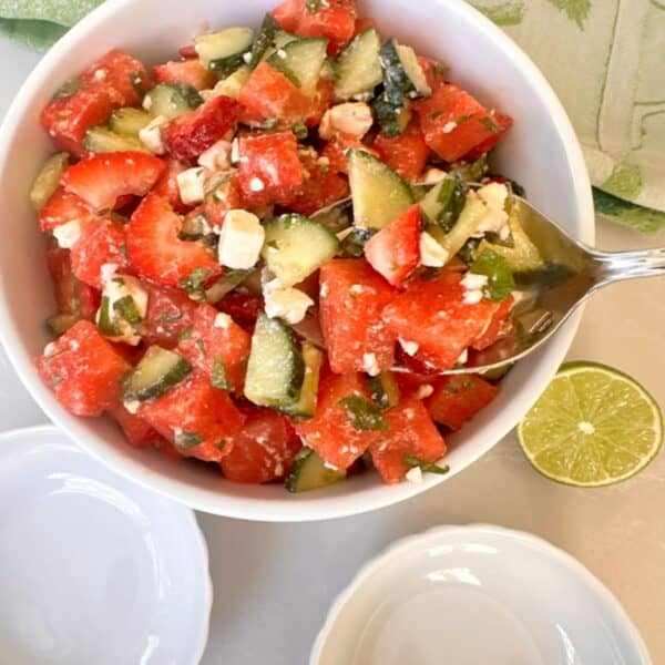 Red, juicy watermelon, strawberry bits, and green-white cucumbers as salad in a white bowl with a large spoon.