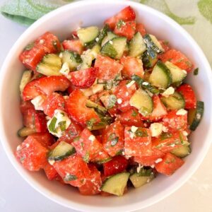White bowl full of juicy red watermelon strawberry cucumber salad with white feta and chopped green mint.