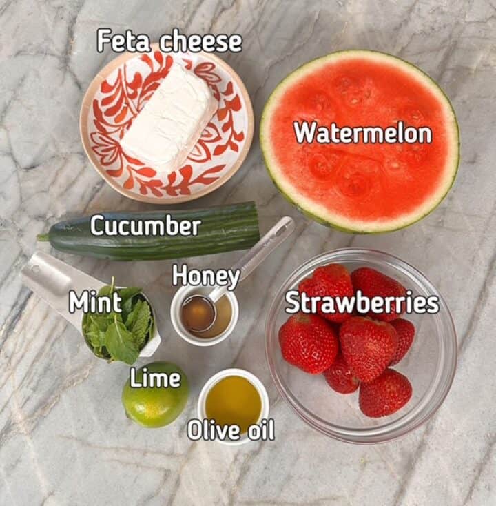 Half a small watermelon, red cut side up, green cucumber, red strawberries, white feta cheese and more for a watermelon salad.