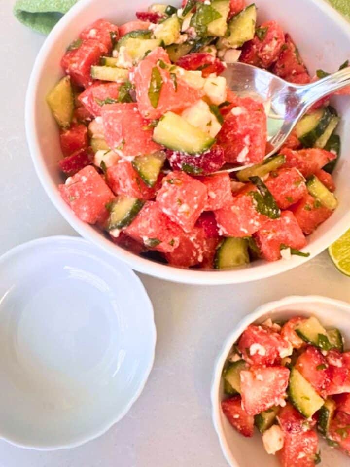 Large white bowl of red and green watermelon strawberry cucumber salad being served into two small white bowls.