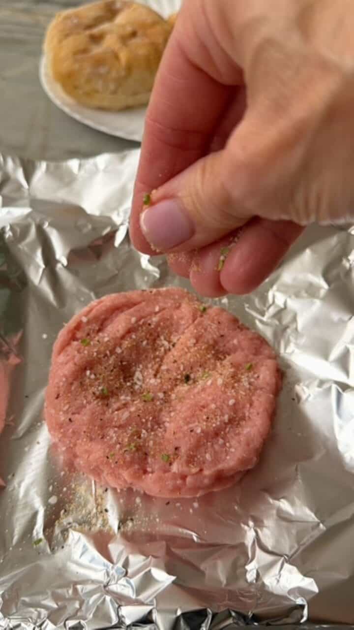 Raw burger patty being seasoned on aluminum foil.