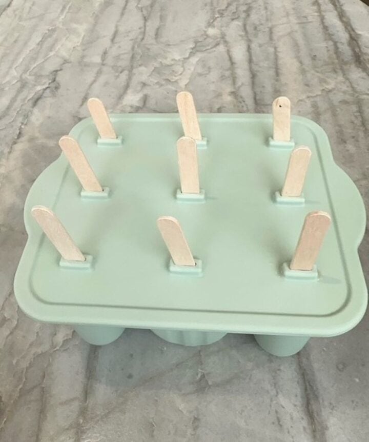 Tan popsicle sticks protrude from a light green silicone popsicle mold.