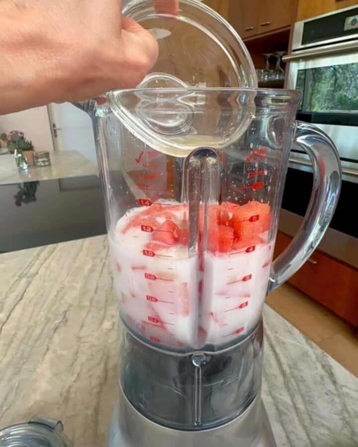 Amber agave nectar is poured into a clear blender container that holds white coconut milk, red watermelon, and lime juice.