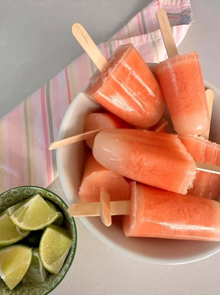 Frozen pink (watermelon) and white-tipped (coconut milk) popsicles in a bowl with green pieces of limes.