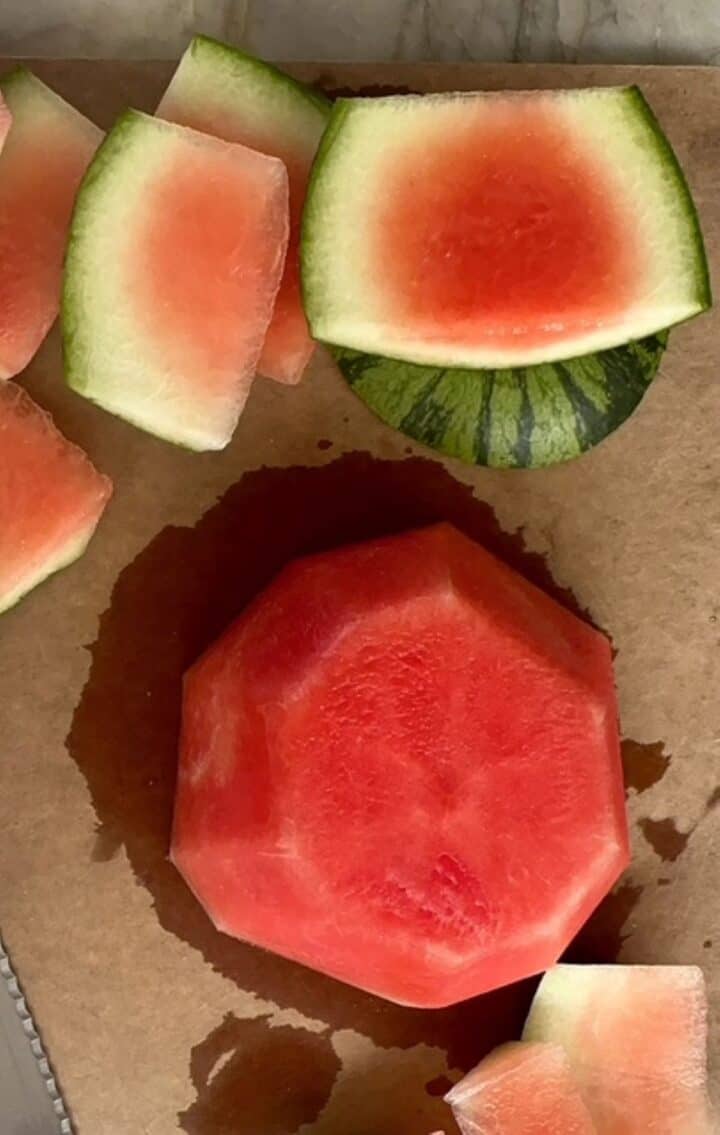 Strips of green and white rind surround a denuded half of a small red watermelon on a brown cutting board.