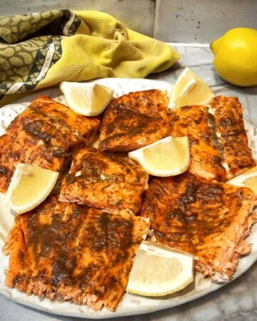 A beige platter loaded with baked Honey-Dijon-Dill glazed salmon fillets, with wedges of yellow lemon as a garnish.