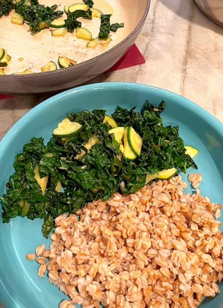 Mound of sauteed green kale and zucchini next to mound of brown farro in a blue shallow bowl.