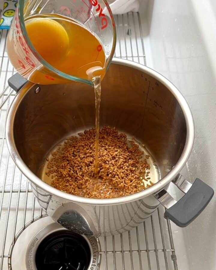 Pouring golden vegetable broth over brown farro in an Instant Pot liner.