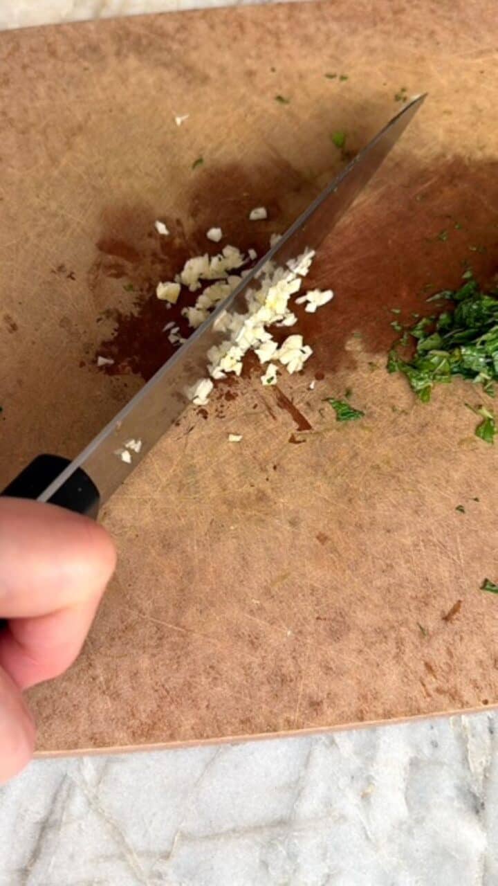 Chef's knife mincing a creamy-colored clove of garlic on a brown cutting board.