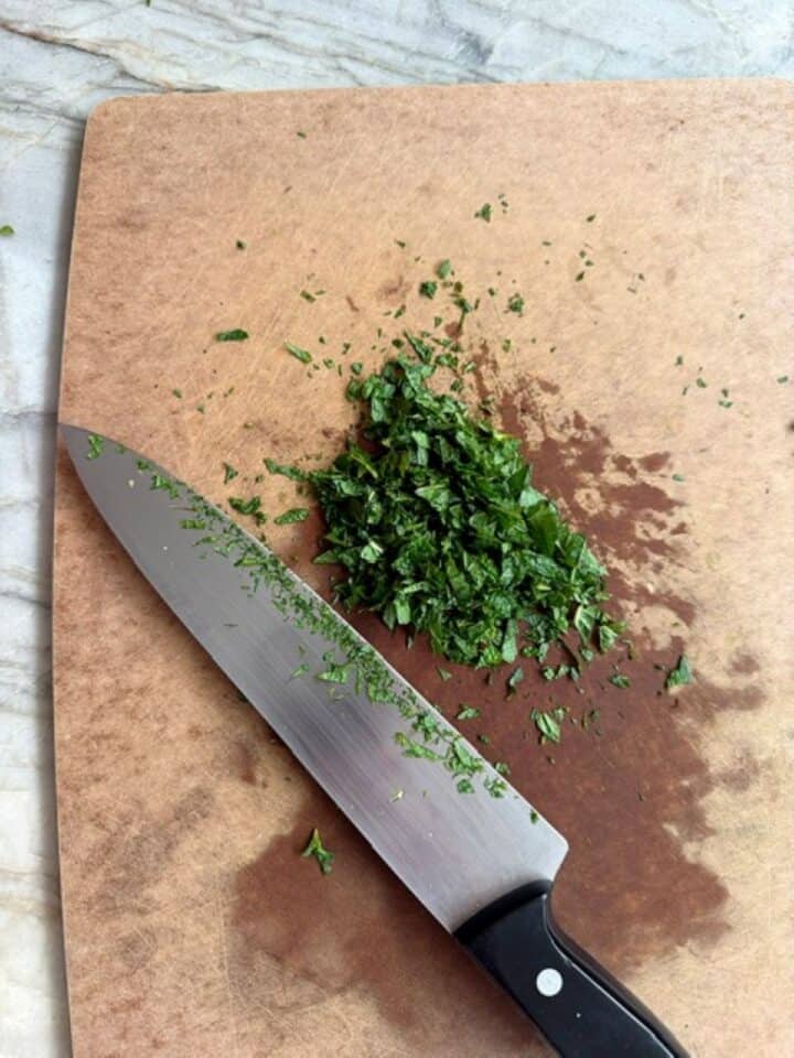 Brown cutting board holding a green pile of minced mint and a chef's knife.