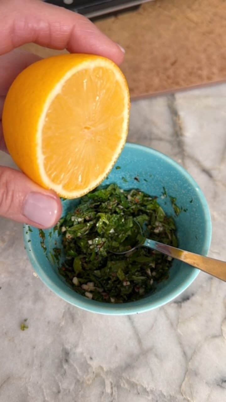 A yellow lemon is squeezed into green mint gremolata in a small blue-green bowl.