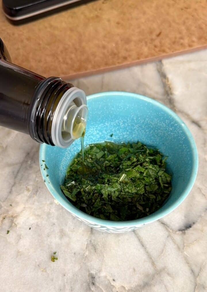Olive oil is poured into minced mint in a small green-blue bowl for gremolata.