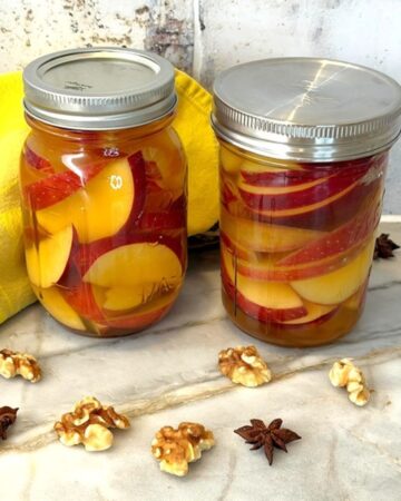 Two jars of pickled apples with spices on a marble surface.