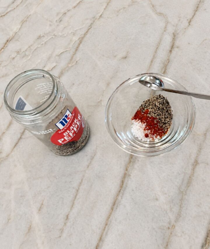 Salt, pepper, and smoked paprika in a bowl with a spoon on marble surface.